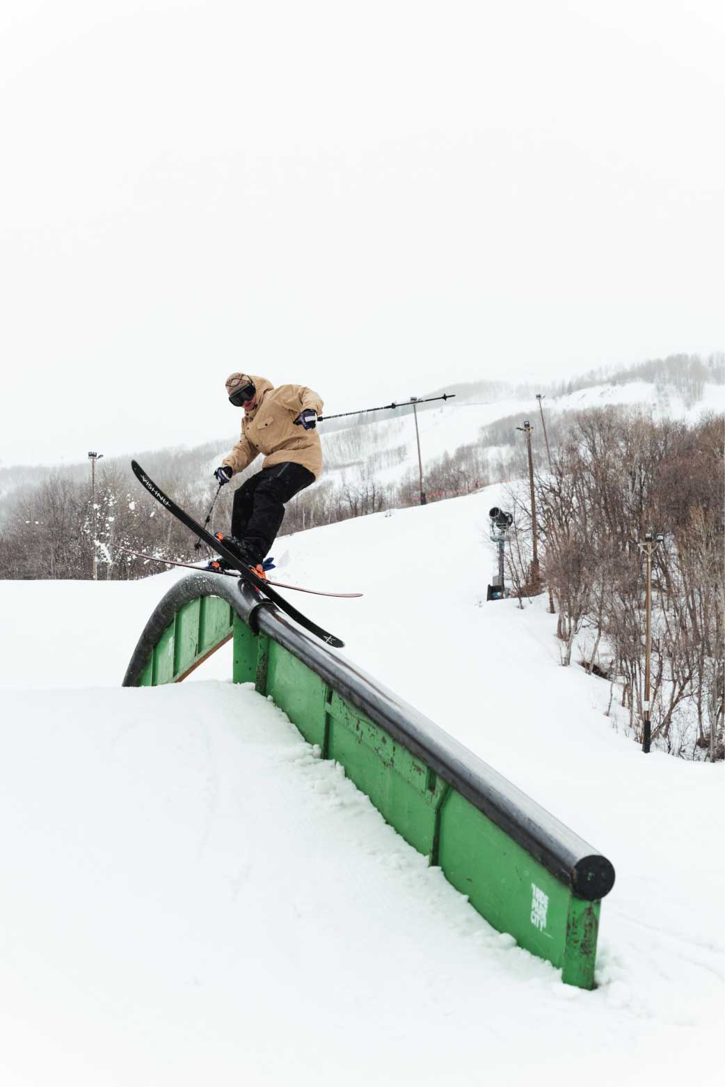 A skier wearing a tan park ski jacket with loose fit slides a rail.