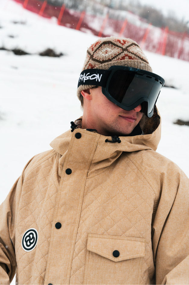 A skier wearing a tan puffer ski jacket with black buttons stands on mountain slope