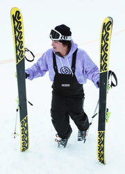 Skier in a black baggy ski bib carrying skis up a mountain slope