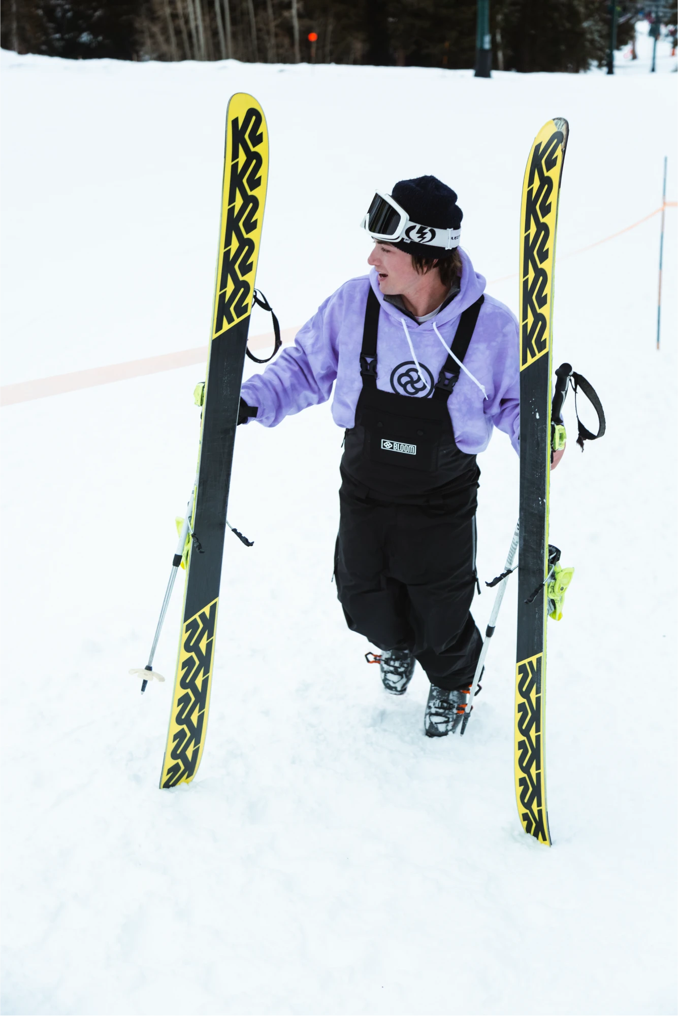 A man wearing black ski bibs hikes up a snowy slope holding his skis and smiling.