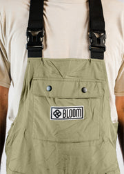 Close-up of a men’s green Bloom Rove snowboard bib showing the chest pocket, raised silicone logo patch, and buckled suspenders.
