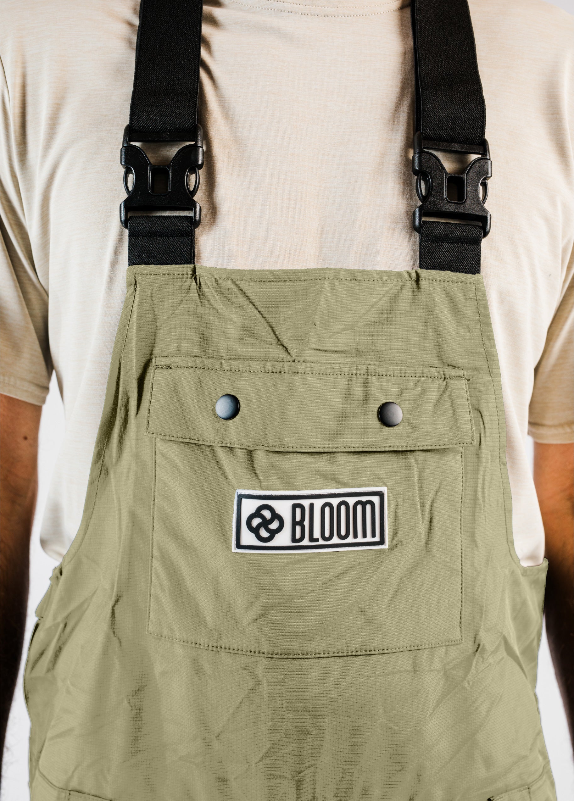 Close-up of a men’s green Bloom Rove snowboard bib showing the chest pocket, raised silicone logo patch, and buckled suspenders.
