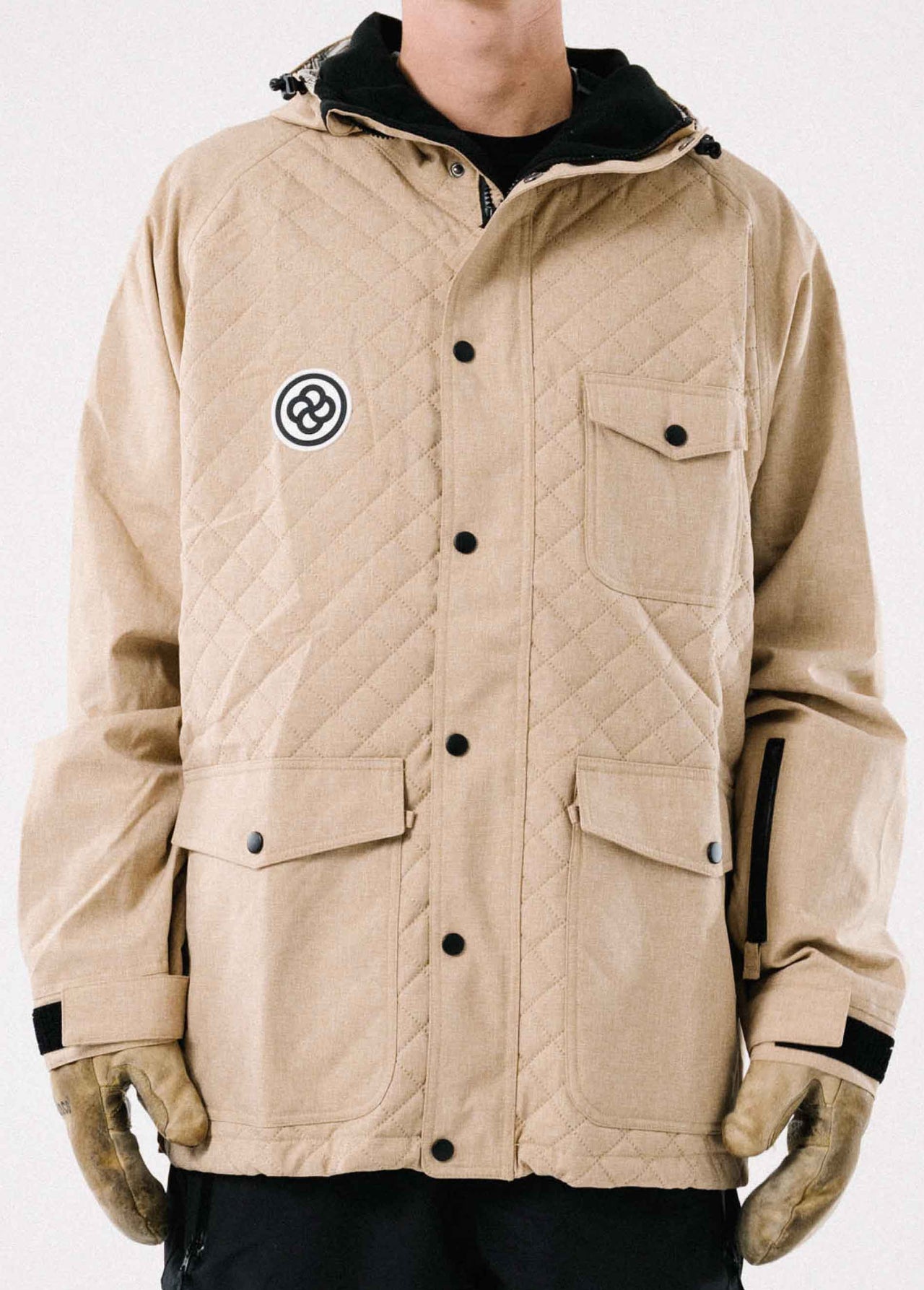 A man wearing a tan ski and snowboard jacket with quilted pattern, lots of pocket, and helmet compatible hood.