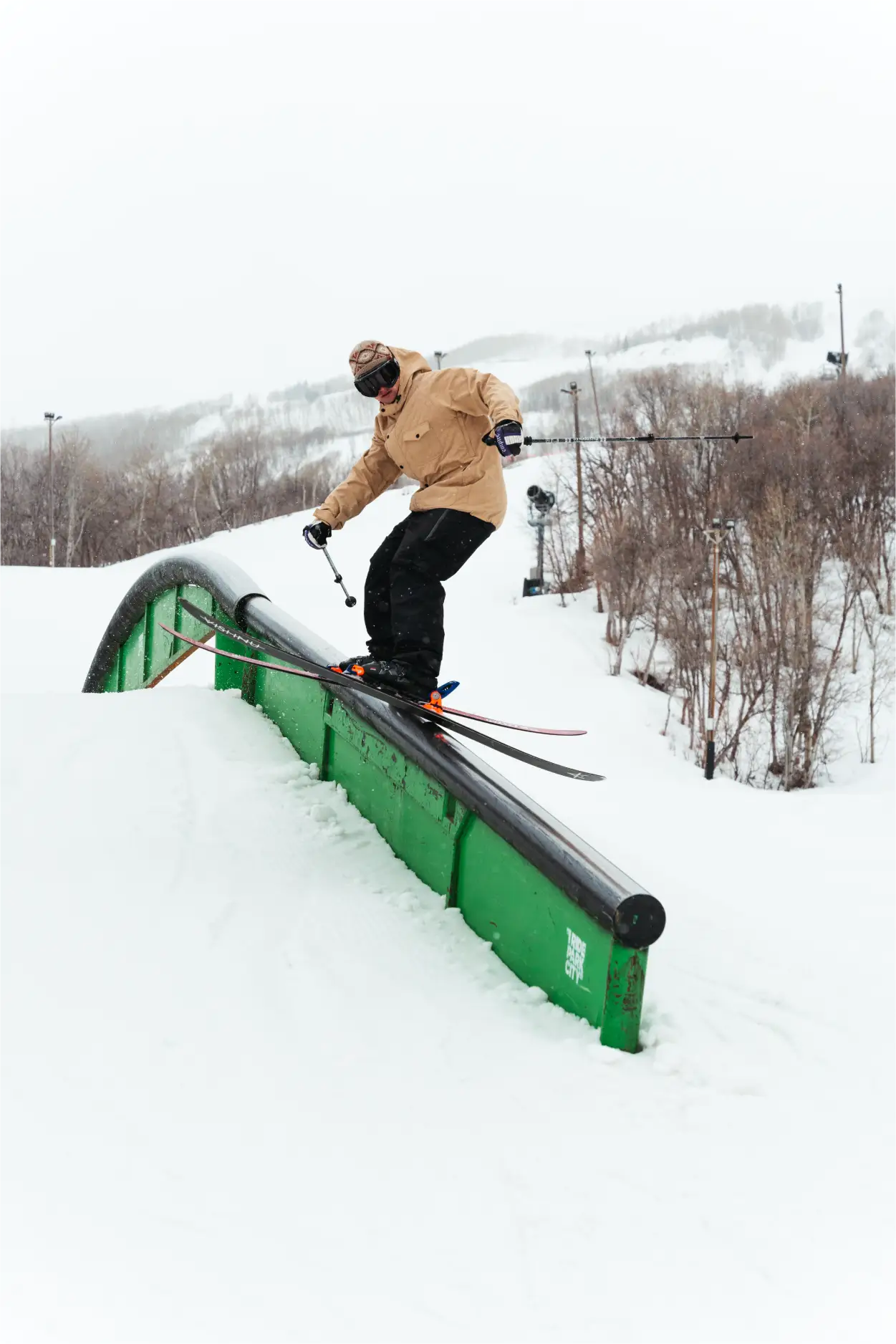 Park skier wearing a tan quilted ski jacket slides a rail