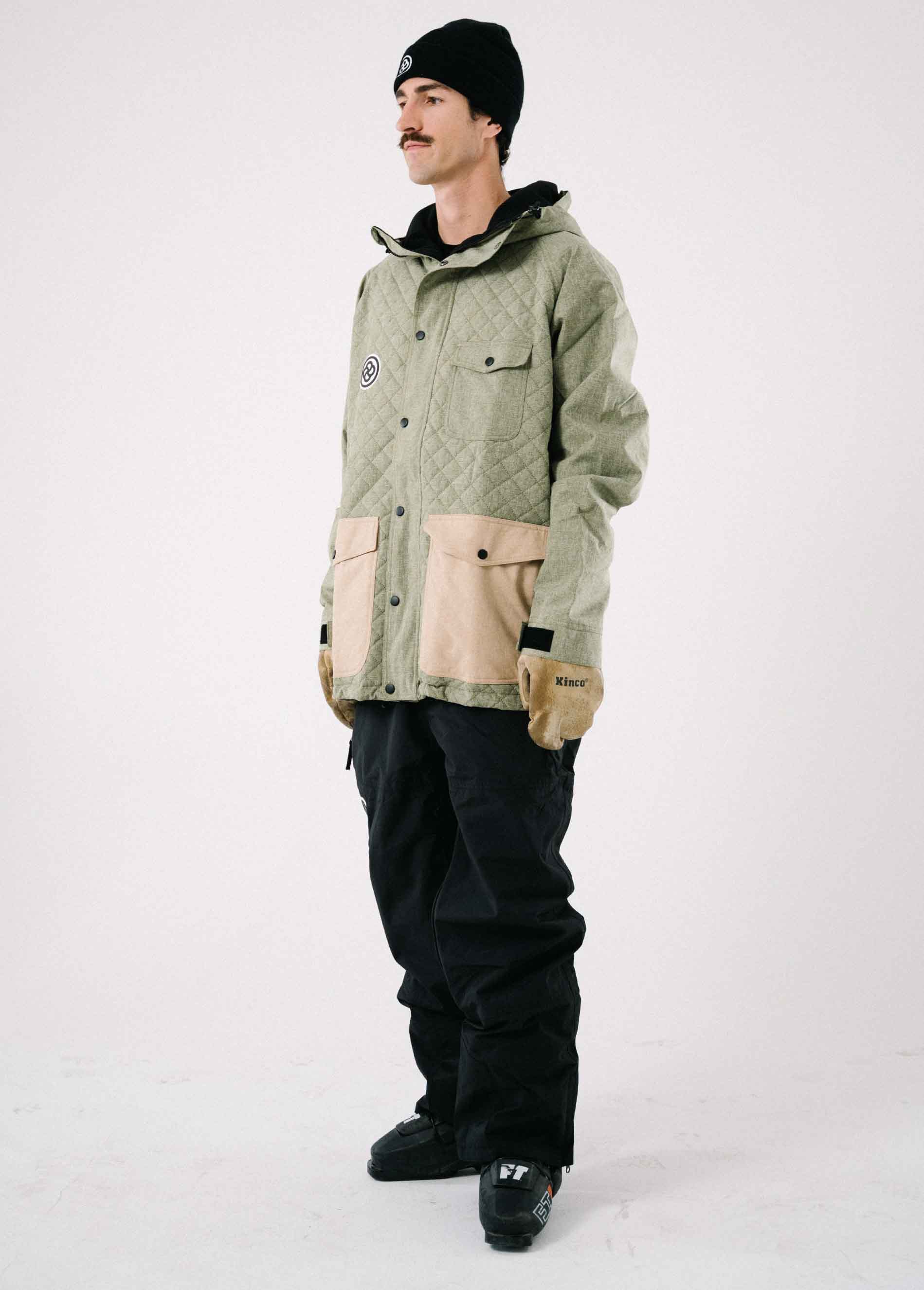 Man wearing a stylish waterproof green ski jacket with black baggy bib pants against a white backdrop